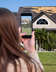 A person with long brown hair, seen from behind, is holding up a smartphone to take a picture of a house with visible roof damage. The phone's screen displays a clear image of the house.