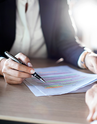 A person in a dark blazer and white shirt is pointing with a silver pen at a document with highlighted sections, suggesting a discussion or review of the text.