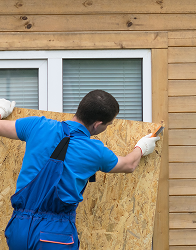 A person in blue overalls and white gloves is boarding up a window with a sheet of plywood on a wooden house, using a utility knife to trim the edges.